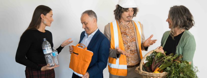 Four people discussing sustainability, holding items like plastic bottles, an orange bag, fresh vegetables, and wearing a construction vest and helmet.